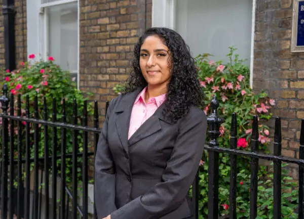 Manusri Prabhakar. Female in grey suit and pink shirt in front of railings and flowers.