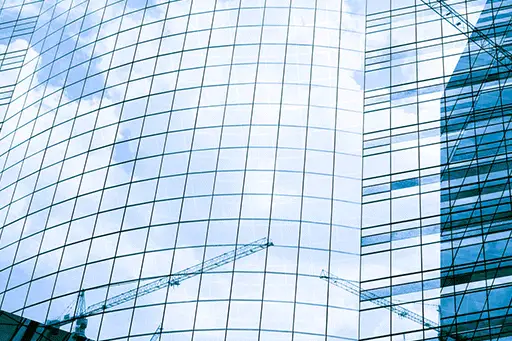 Tall light blue glass office building reflecting some cranes
